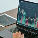Pessoa analisando gráficos de mercado financeiro em laptop com múltiplos monitores e documentos ao lado, focado em investimentos e economia pessoal.