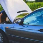 Veículo com o capô aberto na estrada, indicando problema mecânico, com pessoa verificando o motor.