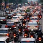 Imagem de trânsito congestionado com carros e motos em uma rua urbana, com luzes de semáforo e prédios ao fundo, ilustrando o fluxo de veículos na cidade.
